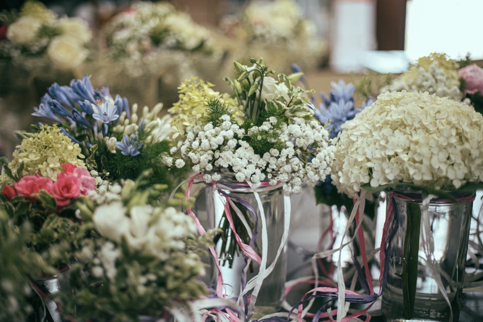 A collection of floral arrangements displayed in tall, clear glass vases, featuring white hydrangeas, delicate white baby's breath, soft pink roses, and blue irises. The flowers are arranged in a lush