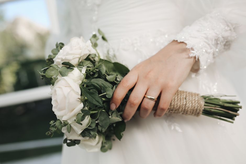 A close-up of a bride's hand holding a bouquet of fresh white roses and eucalyptus leaves, wrapped with twine around the stems. The bride is wearing a white wedding dress with lace and beaded details 