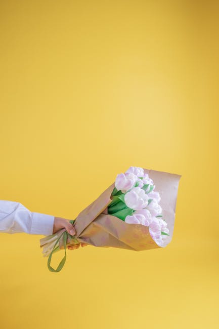 A person's hand, dressed in a white shirt, holds a bouquet of pastel pink tulips wrapped in natural brown paper with a green ribbon against a bright yellow background. The tulips are fresh, with smoot