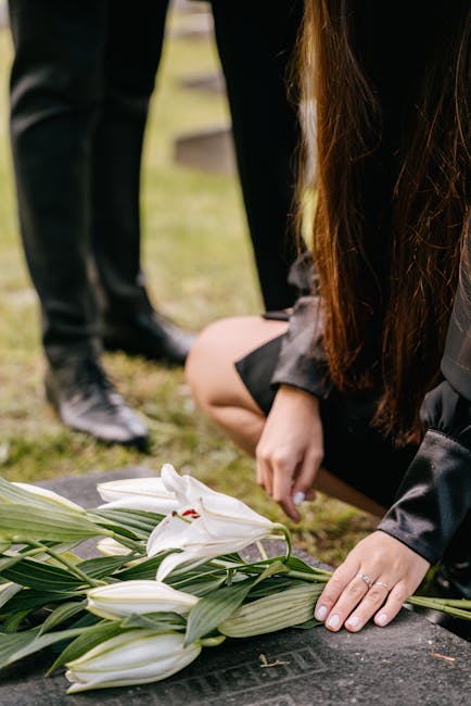 A person with long brown hair, dressed in dark clothing, is kneeling at a grave, gently placing white lilies and greenery on the stone memorial. Another individual, dressed in black, is standing nearb