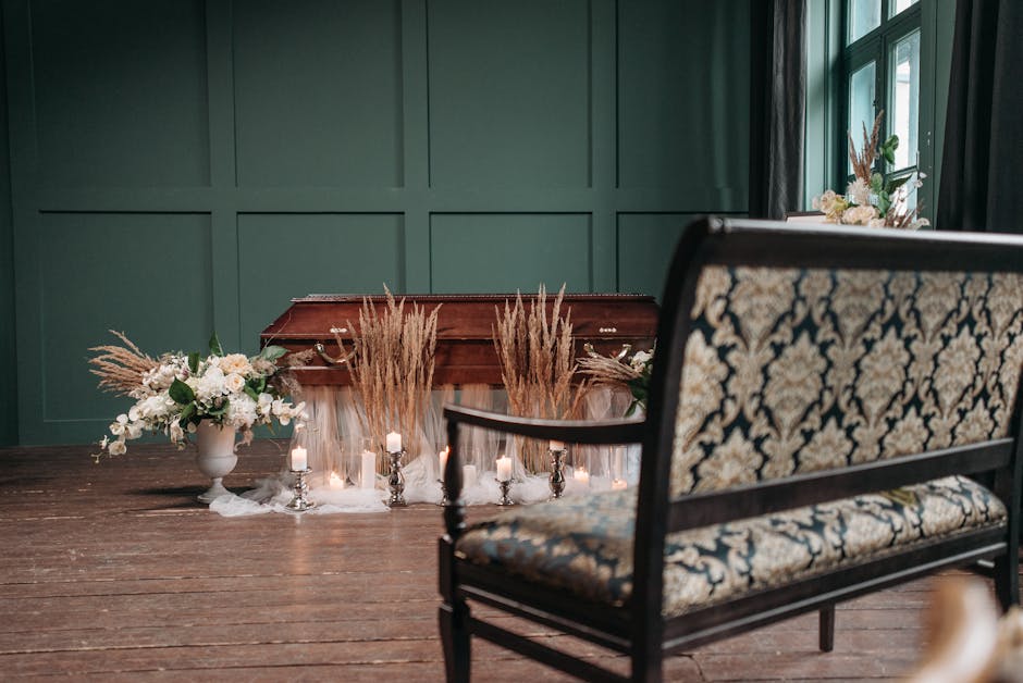 A wooden casket positioned on a dark wooden floor, adorned with arrangements of dried pampas grass and small white candles in silver holders. To the left, a white vase holds a floral bouquet featuring